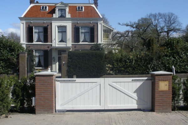 Witte houten poort met diagonale versteviging, gemonteerd tussen bakstenen zuilen bij een klassieke villa met een sierlijke tuin en groene hagen.