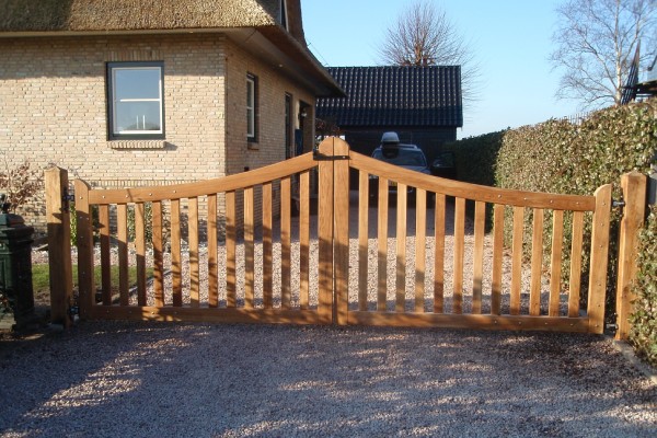 Houten poort met gebogen bovenrand en verticale spijlen, geplaatst bij een oprit met grind en omgeven door een haag, met een bakstenen woning op de achtergrond.